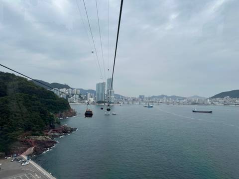 Cable cars glide above the sea toward a modern coastal skyline on a grey day in Busan.