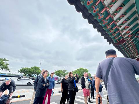 Visitors listen to a guide beneath the painted eaves of a traditional Korean gate on a cloudy day.