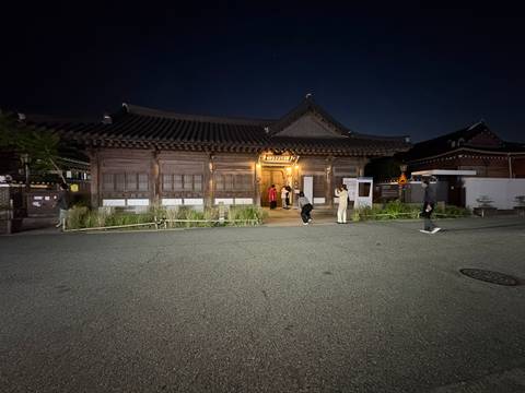 A traditional wooden hanok lit warmly at night as visitors enter the doorway on a quiet street.