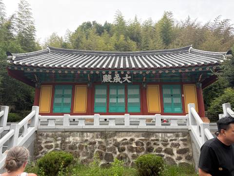 A small, colorful Buddhist shrine with teal doors and yellow walls sits at the edge of a bamboo forest.