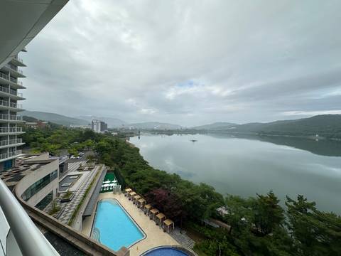 High-angle view from hotel balcony of a calm river or lake surrounded by green mountains and city buildings with an outdoor pool in the foreground.