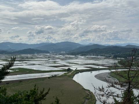 Sweeping view over flooded rice paddies, a winding river and layered mountains under a partly cloudy sky.