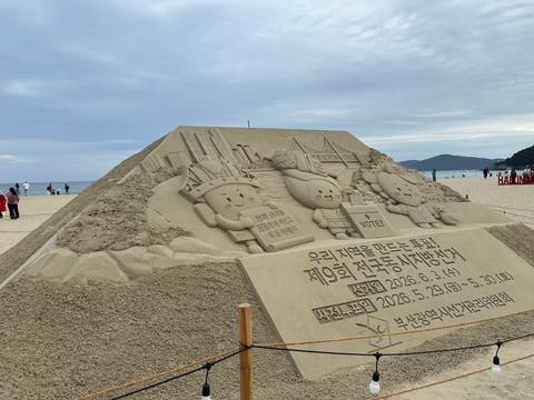 Large sand sculpture on a beach featuring cartoon mascots, skyscrapers and Korean election signage.
