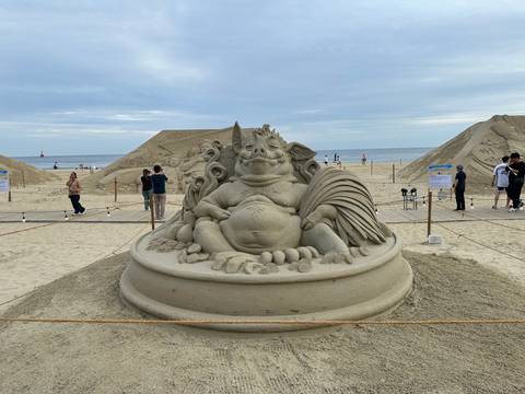 Detailed sand sculpture of a reclining mythic boar surrounded by beachgoers and other sand art displays.