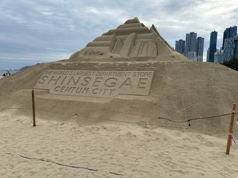 Sand sculpture promoting Shinsegae Centum City with modern skyscrapers in the background.