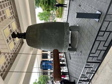 Massive ornate bronze temple bell displayed under a pagoda-style shelter with wooden striker beam.