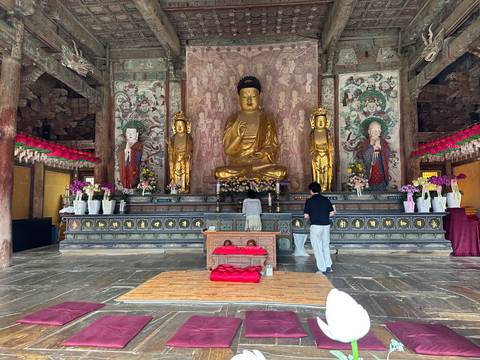 Interior of a Buddhist temple with a golden Buddha altar, flower offerings and two visitors praying on cushions.