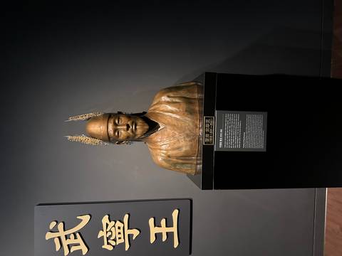 Bronze bust of a historical Korean figure wearing feathered headpieces displayed on a black pedestal in a museum.