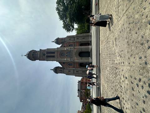 Red-brick Romanesque cathedral with domed towers in bright sunlight as visitors gather on the plaza.
