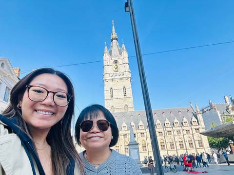 Smiling mother and daughter selfie with the Belfry of Ghent rising behind under a clear blue sky.