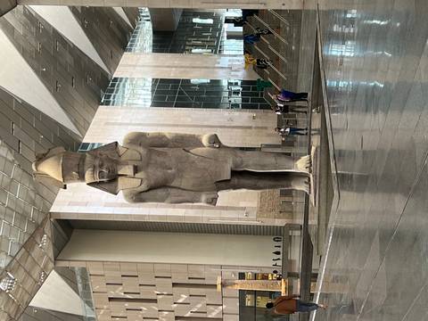 Front view of the towering Ramses II statue inside the Grand Egyptian Museum with a few visitors near its base.