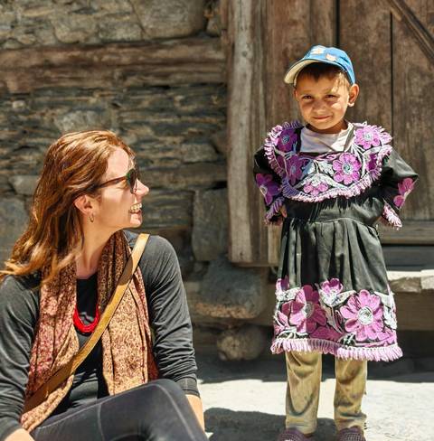 A smiling traveler chats with a young girl in an embroidered dress outside a stone building.