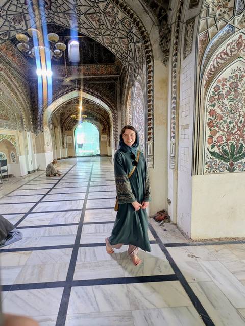 Inside a richly patterned marble mosque corridor, a woman in a green cloak smiles for the camera.