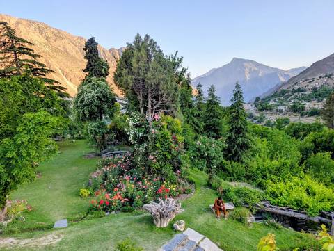 A lush terraced garden bursts with roses and trees against a backdrop of high mountain ridges at sunset.