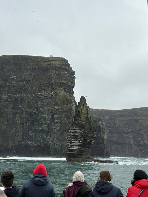 Sheer dark cliffs rise vertically from the Atlantic, dotted with seabirds under an overcast sky.