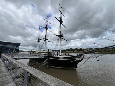 Historic three-masted tall ship docked at a river pier beneath cloudy skies