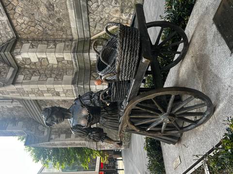 Bronze statue of a woman pushing a cart outside a stone church building