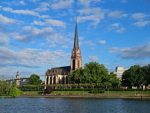 Neo-Gothic church spire rising above leafy riverside park under blue sky in Frankfurt.