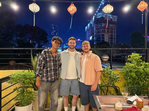 Three friends pose on a rooftop terrace at night with city lights and high-rise construction in the background.