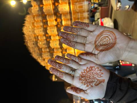 Two hands decorated with fresh henna designs held up against the illuminated façade of Hawa Mahal at night.