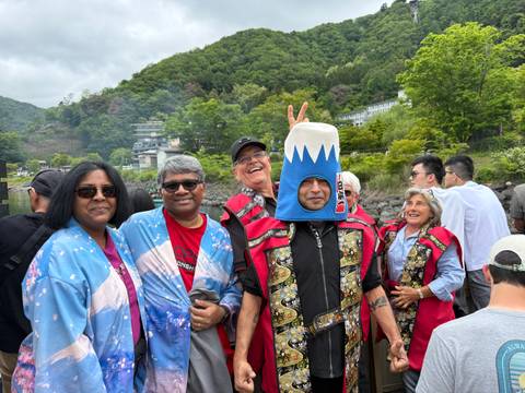 Group of tourists in festive costumes, one wearing a Mount Fuji hat, smiling for camera outdoors