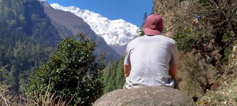 A lone trekker sits on a boulder looking toward snow-covered Himalayan peaks under a clear blue sky.