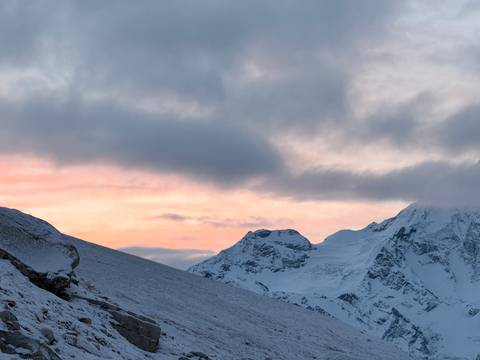 Soft pink sunrise light bathes snow-capped Himalayan peaks beneath dramatic clouds.
