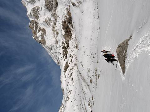 Trekker group with poles makes its way across a vast snowy pass toward jagged Himalayan cliffs.