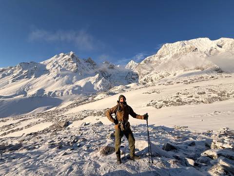 A bundled trekker poses amid a snowy high-altitude plateau ringed by bright white peaks.