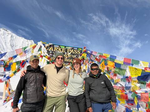 Four trekkers celebrate at Larke Pass sign surrounded by colorful prayer flags and snowfields.