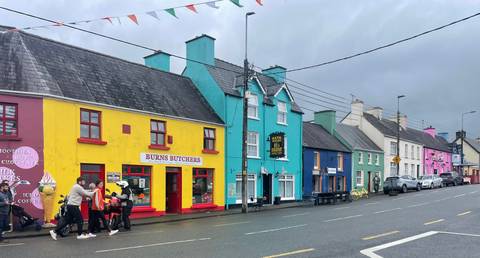 Row of brightly painted village shops on a wet Irish street with pedestrians and strollers.