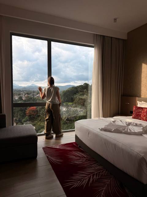 Woman stands by a large hotel window overlooking forested hills and distant mountains.