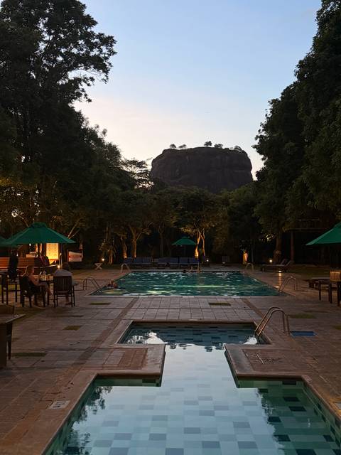 Evening view of a lit swimming pool with Sigiriya Rock silhouetted against the dusk sky.