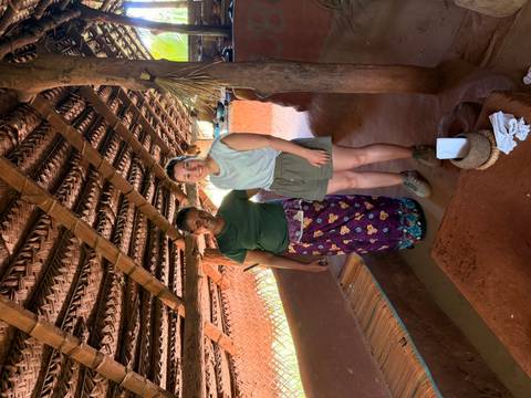 Visitor stands with a local woman inside a rustic thatched kitchen hut.