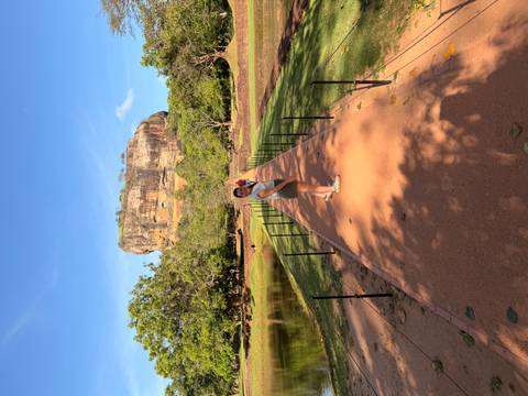 Woman stands on a long path pointing toward Sigiriya Rock rising beyond lush gardens.
