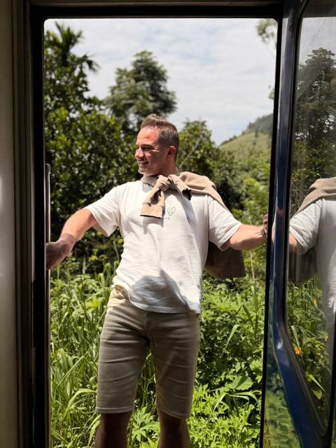 Man smiling at the open door of a train surrounded by lush greenery.