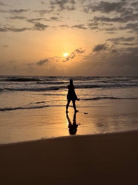 Silhouette of woman walking along wet sand with waves reflecting an orange sunset.