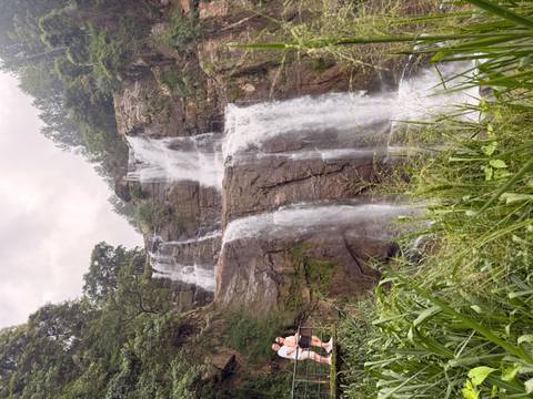 Tall multi-tier waterfall cascades down rock cliffs into lush vegetation with a tiny spectator for scale.