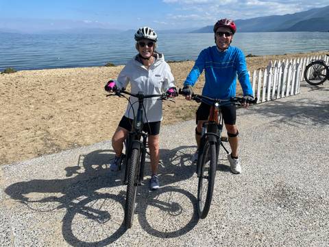Two cyclists pose with e-bikes on a gravel path beside a calm lake.