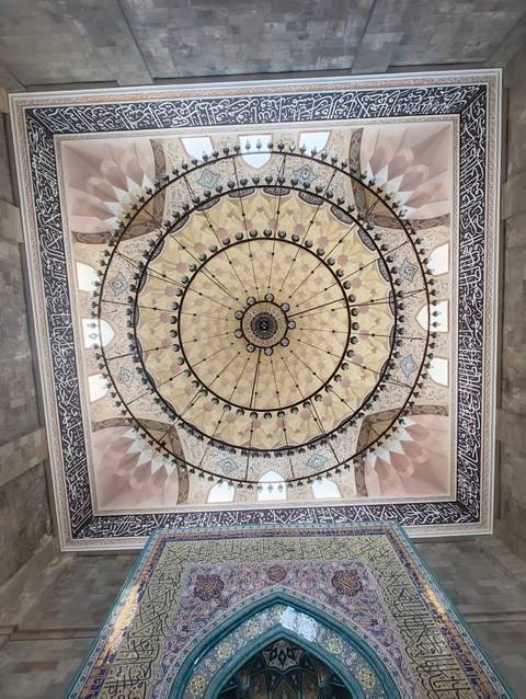 Symmetrical ornate mosque ceiling with concentric floral patterns, chandeliers and Arabic calligraphy.