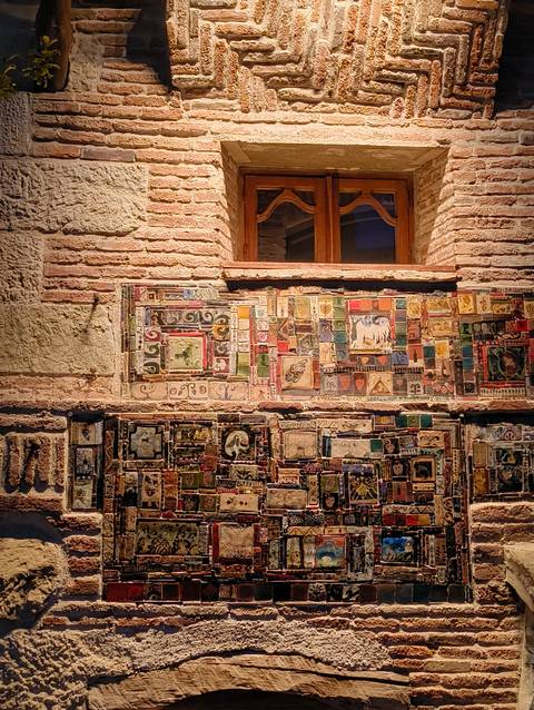 Exterior brick wall adorned with an eclectic mosaic of colorful ceramic tiles and artwork beneath a wooden window.