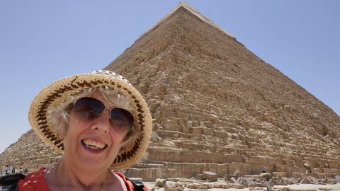 Woman in sunhat smiles in front of a massive limestone pyramid under clear desert sky.