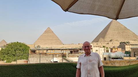 Man standing on a rooftop terrace with the Great Pyramids visible beyond city buildings and greenery.