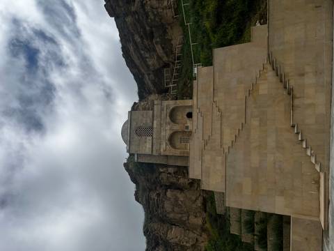 Tall beige stone mausoleum built into a cliff face viewed from below under cloudy skies.
