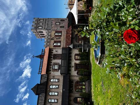 Whimsical castle-style winery building with small fountain and bright red rose in the foreground against a partly cloudy blue sky.