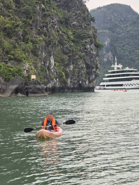 Man in kayak paddling through emerald waters with towering limestone cliffs and cruise ship behind.