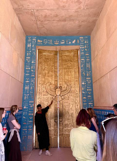 People posing before large ornate golden doors decorated with hieroglyph motifs.