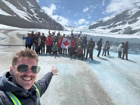 Large tour group poses on a blue glacial ice sheet with Canadian flag while mountains rise behind