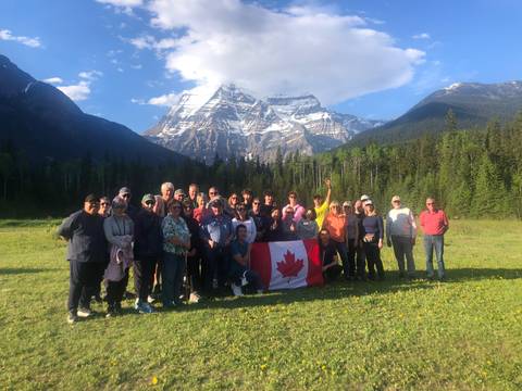 Tour group proudly holding a Canadian flag in a green meadow with towering Mount Robson behind.