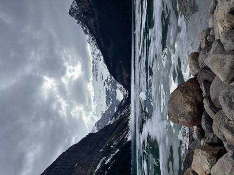 Part-frozen Lake Louise lined with boulders, ringed by snowy peaks under a moody cloudy sky.